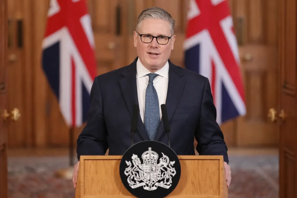 Starmer Says US Can Use UK Bases for Defence UK Prime Minister Keir Starmer delivering a statement from a podium with Union Jack flags in the background regarding the use of UK bases amid Iran tensions.